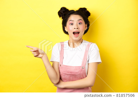 Excited asian teen girl pointing finger left at logo and looking happy, showing advertisement, standing on yellow background Excited asian teen girl pointing finger left at logo and looking happy, showing advertisement, standing on yellow background 81857506
