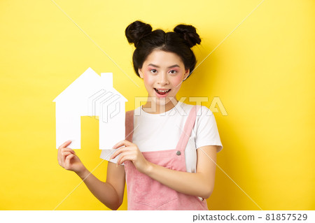 Real estate and family concept. Cute asian woman with bright makeup and stylish hairbuns, showing paper house cutout, smiling determined, yellow background Real estate and family concept. Cute asian woman with bright makeup and stylish hairbuns, showing paper house cutout, smiling determined, yellow background 81857529