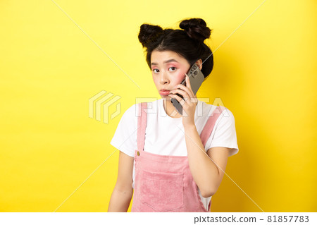 Cute teen girl talking on smartphone, making silly pouting face and look timid at camera, standing with glamour makeup on yellow background Cute teen girl talking on smartphone, making silly pouting face and look timid at camera, standing with glamour makeup on yellow background 81857783