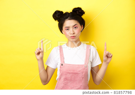 Annoyed and tired asian girl with pink glamour makeup, pointing fingers up and look bothered, standing on yellow background 81857798