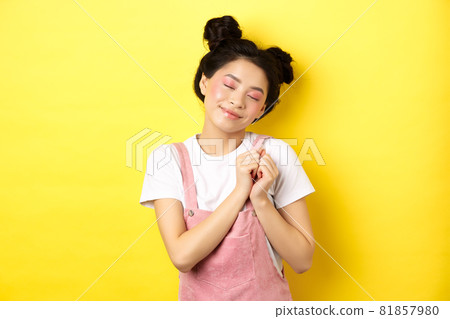 Cute asian girl with makeup, close eyes and remember beautiful moment, holding hands on heart daydreaming, standing on yellow background 81857980