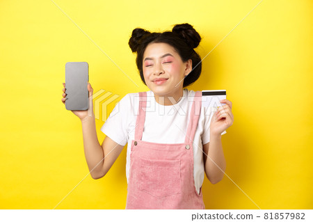 Online shopping concept. Happy asian girl showing empty smartphone screen and credit card, paying contactless, standing on yellow background 81857982