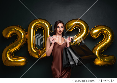 Woman in elegant dress hold credit bank card and shopping bags on black background, numbers air balloons studio. 81862012