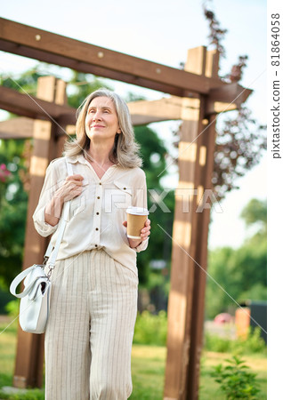 Calm smiling woman with coffee on walk in park Calm smiling woman with coffee on walk in park 81864058