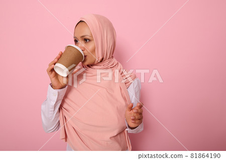 Beautiful Arabic muslim woman with covered head drinking hot drink, tea or coffee from disposable cardboard takeaway cup, standing sideways, three quarters, against pink background with copy space 81864190