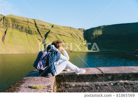 Back view woman with backpack sitting near mountain's lake and enjoying the view of green hills - her hike destination. Travel lifestyle wanderlust adventure. Vacation in nature. Freedom and calm Back view woman with backpack sitting near mountain's lake and enjoying the view of green hills - her hike destination. Travel lifestyle wanderlust adventure. Vacation in nature. Freedom and calm 81865569