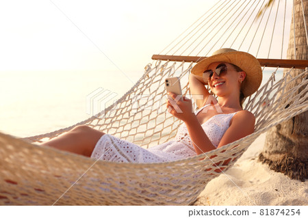 Working at sunset. Young happy female using smartphone while lying in the hammock on the beach 81874254