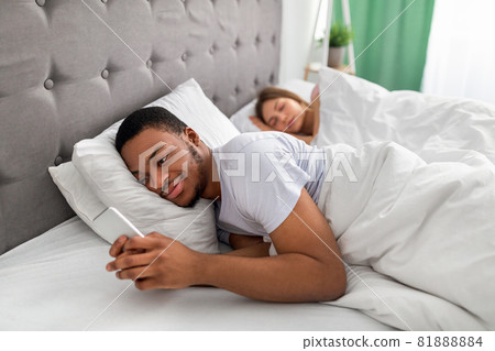 Young black guy lying on bed and using mobile phone to text his lover while his wife is asleep. Relationship infidelity Young black guy lying on bed and using mobile phone to text his lover while his wife is asleep. Relationship infidelity 81888884