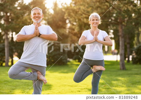 Positive senior couple performing yoga on green lawn in park during sunrise 81889248