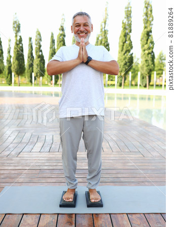 Calm senior yogi man standing barefoot on bed of nails with hands in namaste gesture outdoors Calm senior yogi man standing barefoot on bed of nails with hands in namaste gesture outdoors 81889264