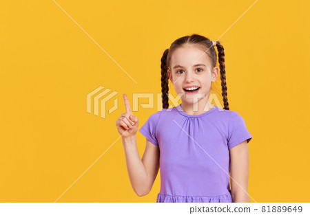 Happy kid girl pointing up while standing in yellow studio Happy kid girl pointing up while standing in yellow studio 81889649