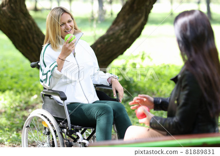 Disabled woman in wheelchair eating apple with her friend in park Disabled woman in wheelchair eating apple with her friend in park 81889831