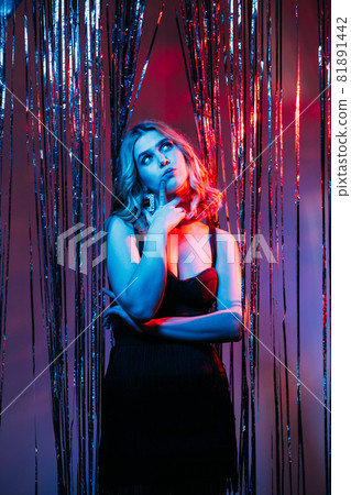 Christmas party. Puzzled woman. Find decision. Festive look. Thoughtful lady black dress curly hair looking up choose something in shimmering silver cascade curtain blue red neon light. 81891442