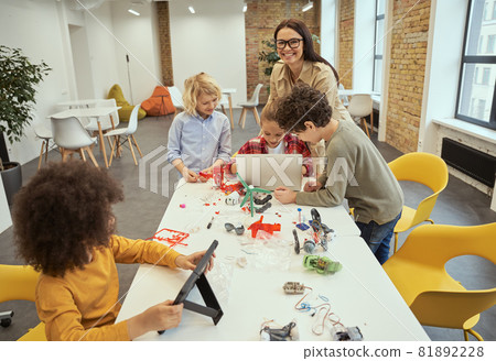 Happy young female teacher in glasses smiling at camera while helping kids to build robotic toys and programming them using computer during STEM class 81892228