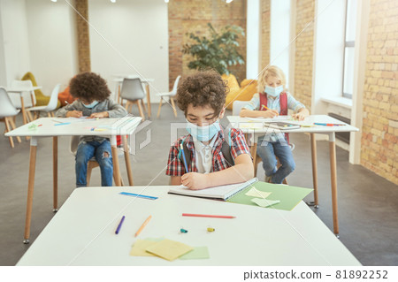 School boy wearing face mask during coronavirus outbreak writing in his notebook while sitting at the desk in classroom. Kids studying in elementary school 81892252