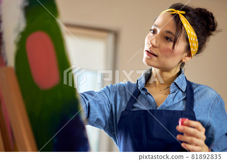 Portrait of young woman in apron looking inspired while working on abstract painting at cozy workshop 81892835