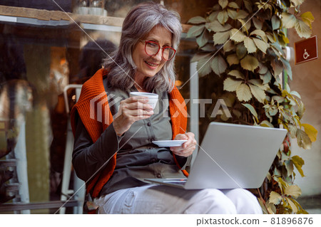 Pretty mature woman with cup at videochat via laptop sits on bench near cafe outdoors Pretty mature woman with cup at videochat via laptop sits on bench near cafe outdoors 81896876