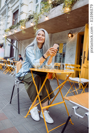 Thoughtful senior Asian womanr with phone sits at small table on outdoors cafe terrace Thoughtful senior Asian womanr with phone sits at small table on outdoors cafe terrace 81896879