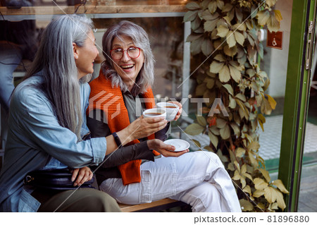 Senior Asian woman with friend clink cups of drinks sitting on bench near cafe outdoors Senior Asian woman with friend clink cups of drinks sitting on bench near cafe outdoors 81896880