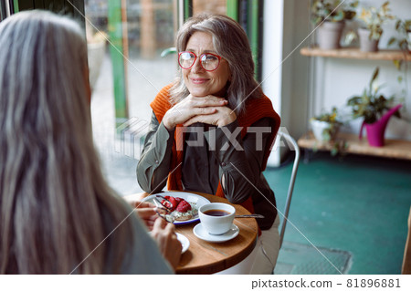 Grey haired woman with glasses looks at friend sitting at small table in cozy cafe Grey haired woman with glasses looks at friend sitting at small table in cozy cafe 81896881