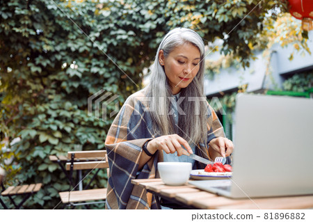 Positive mature Asian woman eats dessert in front of laptop at table on outdoors cafe terrace Positive mature Asian woman eats dessert in front of laptop at table on outdoors cafe terrace 81896882