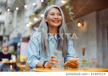 Cheerful mature Asian woman with glass of water and phone sits at small table outdoors 81897014