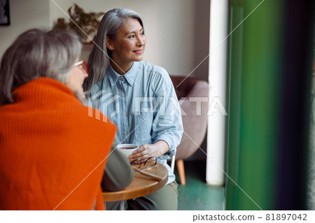 Cheerful senior Asian woman with friend look out of window at table in cozy cafe Cheerful senior Asian woman with friend look out of window at table in cozy cafe 81897042