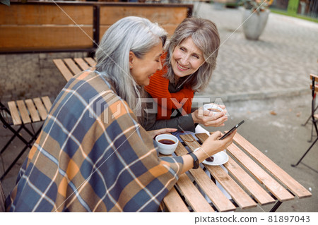 Mature Asian woman shows mobile phone to best friend in street cafe on autumn day Mature Asian woman shows mobile phone to best friend in street cafe on autumn day 81897043