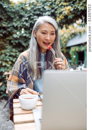 Beautiful senior Asian woman eats fresh strawberry in front of laptop at table outdoors Beautiful senior Asian woman eats fresh strawberry in front of laptop at table outdoors 81897053