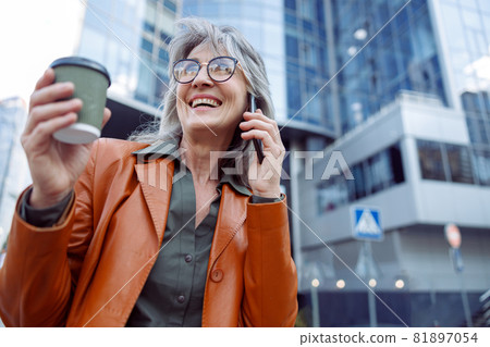 Happy female pensioner with cup of drink n talks on mobile phone on modern city street Happy female pensioner with cup of drink n talks on mobile phone on modern city street 81897054