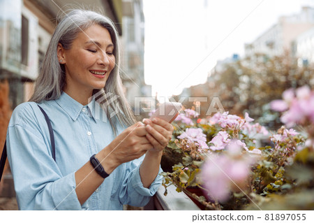 Smiling grey haired Asian woman uses cellphone standing on outdoors terrace Smiling grey haired Asian woman uses cellphone standing on outdoors terrace 81897055