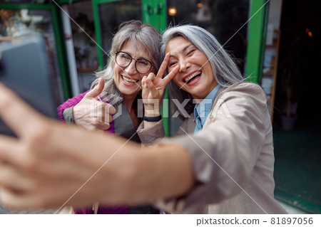 Attractive mature Asian woman with friend take selfie gesturing with phone on city street Attractive mature Asian woman with friend take selfie gesturing with phone on city street 81897056