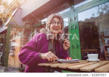 Smiling senior woman in knitted jacket eats tasty strawberry dessert at table in outdoors cafe Smiling senior woman in knitted jacket eats tasty strawberry dessert at table in outdoors cafe 81897057