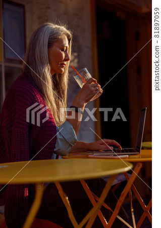 Senior Asian woman holds glass sitting in front of laptop at table on cafe terrace Senior Asian woman holds glass sitting in front of laptop at table on cafe terrace 81897059
