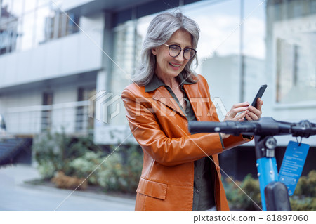 Smiling mature woman uses phone standing near e-scooter exposed on city street Smiling mature woman uses phone standing near e-scooter exposed on city street 81897060