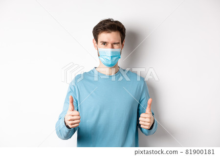 Covid-19, pandemic and social distancing concept. Happy young man in medical mask winking, showing thumbs up in approval, recommending product, white background Covid-19, pandemic and social distancing concept. Happy young man in medical mask winking, showing thumbs up in approval, recommending product, white background 81900181