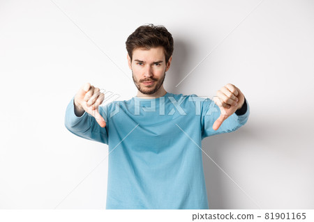 Skeptical handsome guy with beard, showing thumbs down in disapproval, dislike something bad, standing over white background 81901165