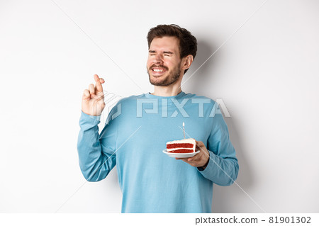 Celebration and holidays concept. Young man celebrating birthday, cross fingers for good luck, making wish on bday cake with lit candle, standing over white background Celebration and holidays concept. Young man celebrating birthday, cross fingers for good luck, making wish on bday cake with lit candle, standing over white background 81901302