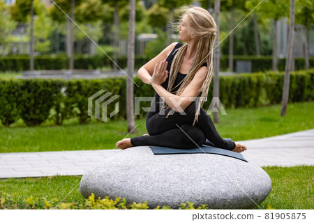Young beautiful woman with blonde hair doing yoga exercise in green public park in summer morning, outdoors. Healthy lifestyle, mental health concept. 81905875