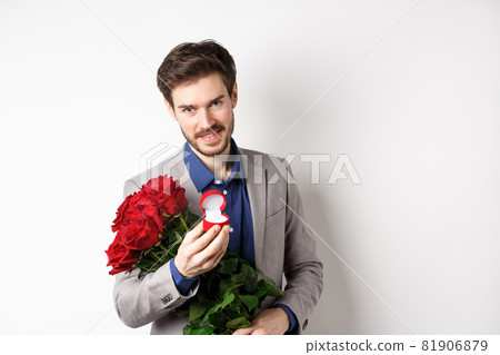 Romantic man with boquet of red roses asking to marry him, holding engagement ring and looking confident at camera, standing in suit over white background 81906879