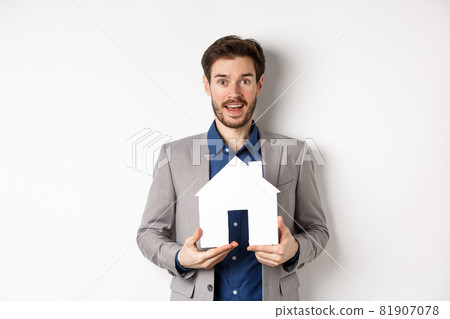 Real estate. Excited male entrepreneur holding paper house cutout, wearing suit and look hopeful at camera 81907078