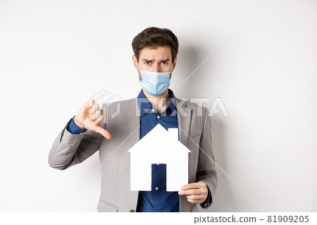 Real estate and covid-19 concept. Disappointed guy in medical mask and suit showing paper house cutout with thumbs-down, complaining on agency, white background Real estate and covid-19 concept. Disappointed guy in medical mask and suit showing paper house cutout with thumbs-down, complaining on agency, white background 81909205