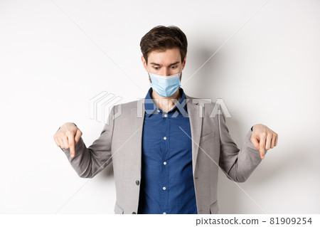 Covid-19, pandemic and business concept. Surprised guy in medical mask and suit looking and pointing down with popped eyes, gasping amazed, white background Covid-19, pandemic and business concept. Surprised guy in medical mask and suit looking and pointing down with popped eyes, gasping amazed, white background 81909254