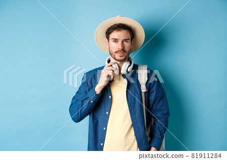 Handsome guy travelling with headphone, wearing straw hat and smiling, standing with backpack on blue background Handsome guy travelling with headphone, wearing straw hat and smiling, standing with backpack on blue background 81911284