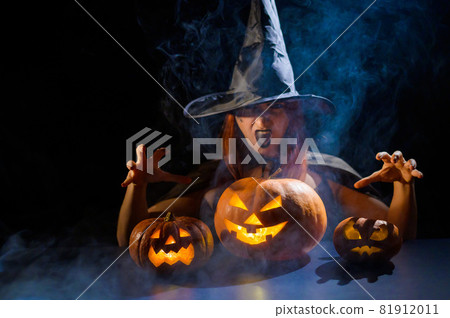The evil witch casts a spell on pumpkins. Portrait of a woman in a carnival halloween costume in the dark The evil witch casts a spell on pumpkins. Portrait of a woman in a carnival halloween costume in the dark 81912011