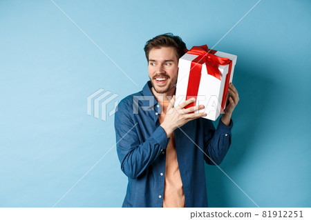 Valentines day. Smiling man guessing what inside gift box, got present from lover girlfriend, standing against blue background 81912251