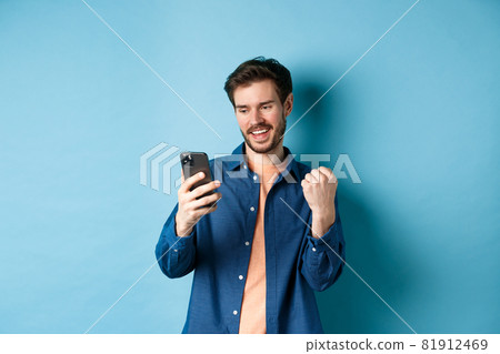 Image of lucky guy winning online, reading mobile phone screen and making fist pump, triumphing, standing on blue background Image of lucky guy winning online, reading mobile phone screen and making fist pump, triumphing, standing on blue background 81912469
