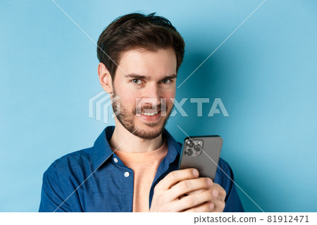 Close-up of attractive caucasian man holding mobile phone and smiling at camera, standing in casual outfit on blue background 81912471