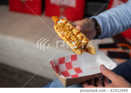 hand holds fried sausage in dough with ketchup in paper red white packaging on the street. hand holds fried sausage in dough with ketchup in paper red white packaging on the street. 81912840