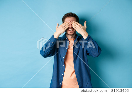 Happy smiling guy waiting for surprise, covering eyes with hands and anticipating gift, standing on blue background 81913493